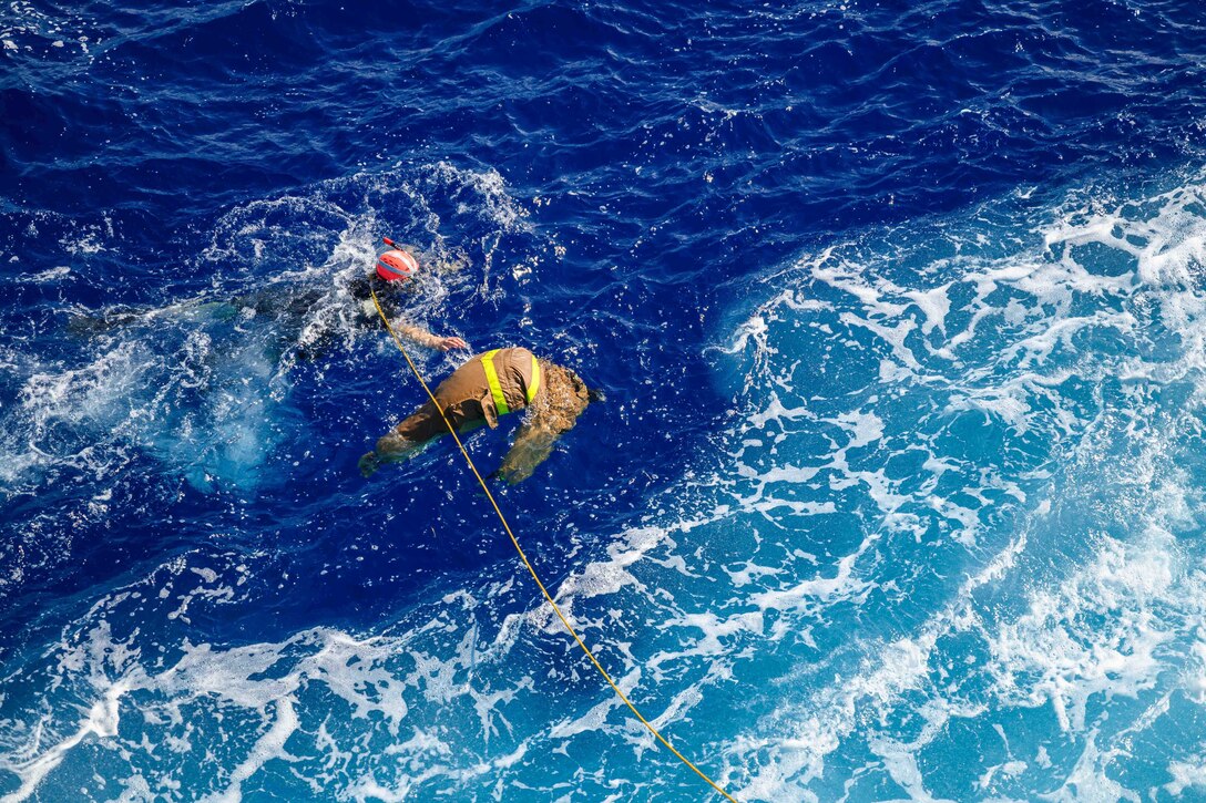 A sailor hoisted to a ship by a yellow rope swims near a simulated casualty in a body of water.