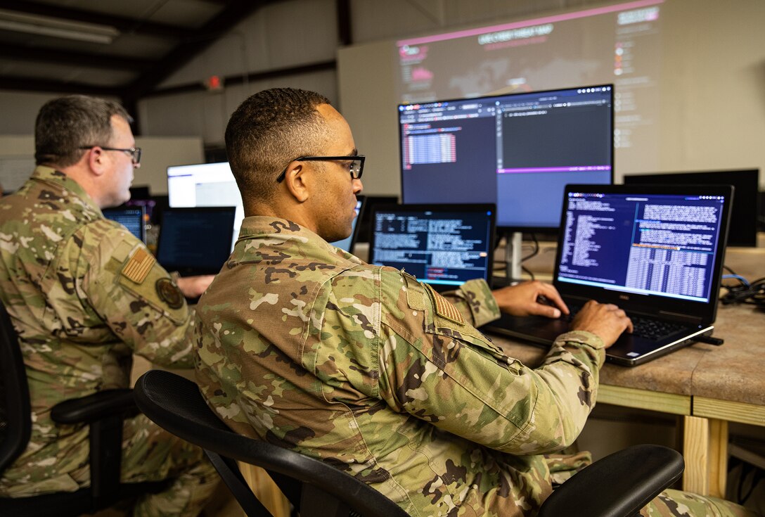 Service members in uniform are seated at computer workstations.