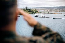 A Marine, shown from behind in blurry foreground, salutes toward a white structure in the sea.
