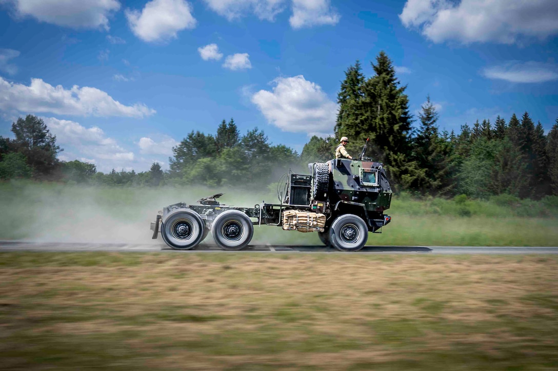 A soldier stands in the cab of a large military vehicle as it moves along a paved road in a rural area.