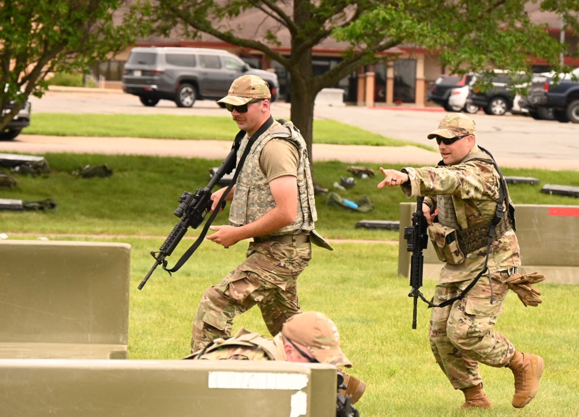 Two males in uniform walk in grass around blockades with simulated weapons.