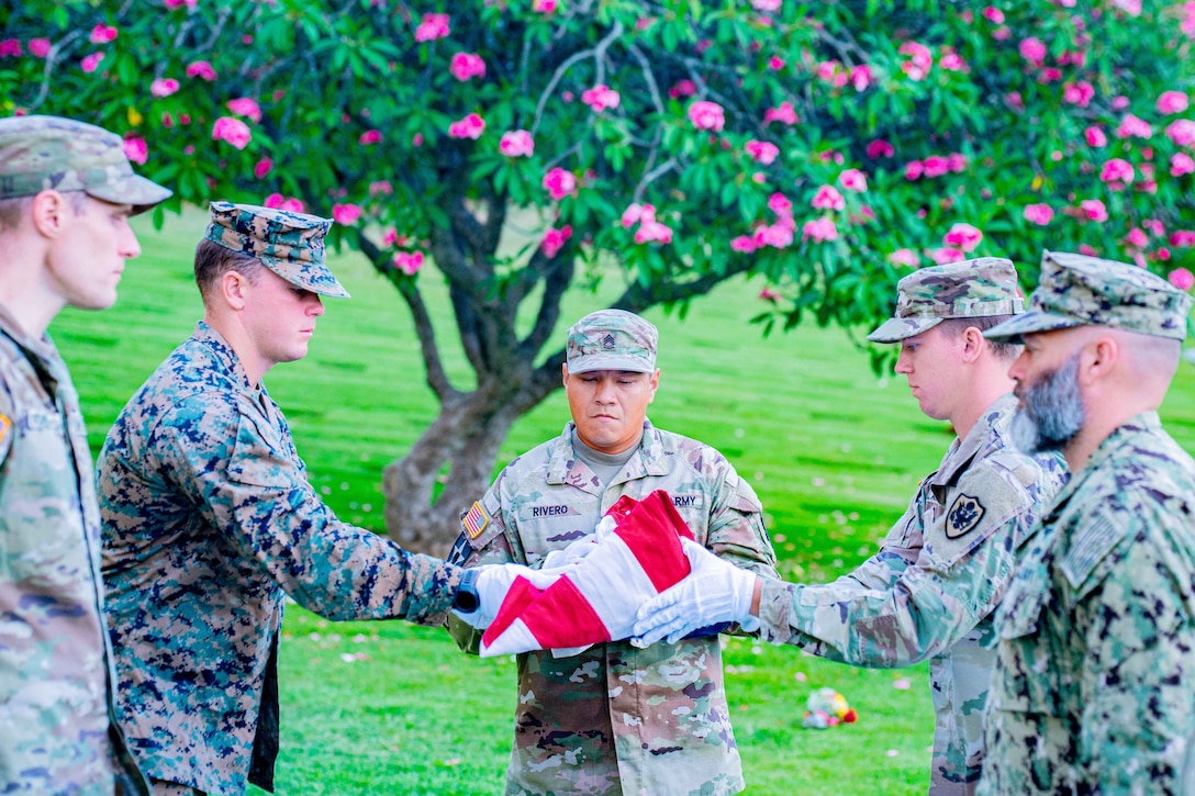 Two service members watch as three others wearing white gloves on their hands unfold an American flag in front of a tree covered in pink flowers.