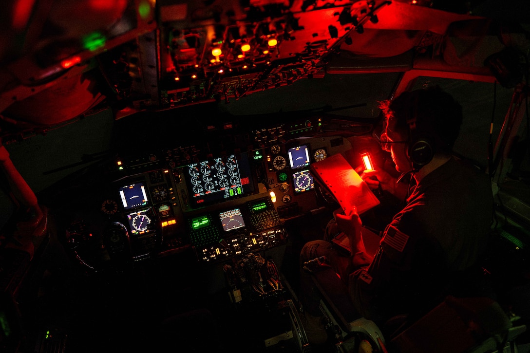 An airman holds a flashlight while reading a notebook in a cockpit illuminated by red, green, white and orange lights.