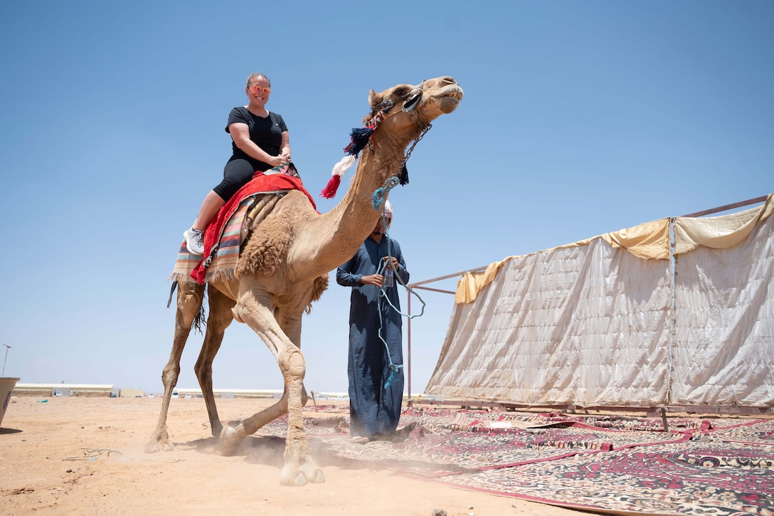 An airman smiles while riding a camel in a desert-like area as a guide holds its reigns.