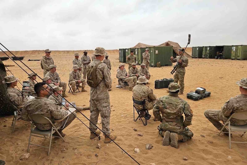 A service member holds a long, thin gadget while talking to a group of service members.