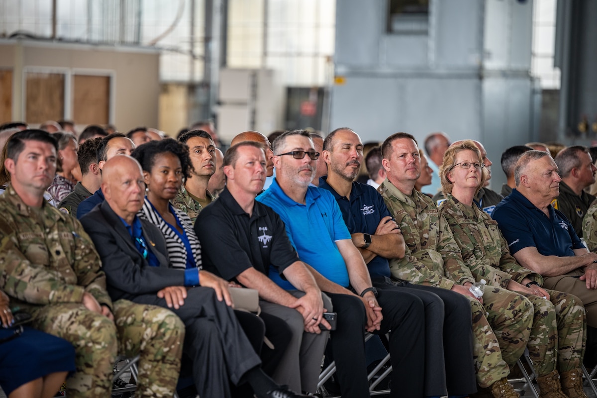 Individuals sit in folding chairs and watch speeches during a ceremony for the F-16 50th anniversary.