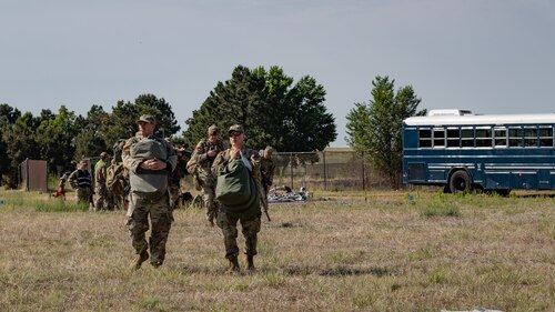 Servicemembers carrying bags walking in a field with a blue bus in the background.
