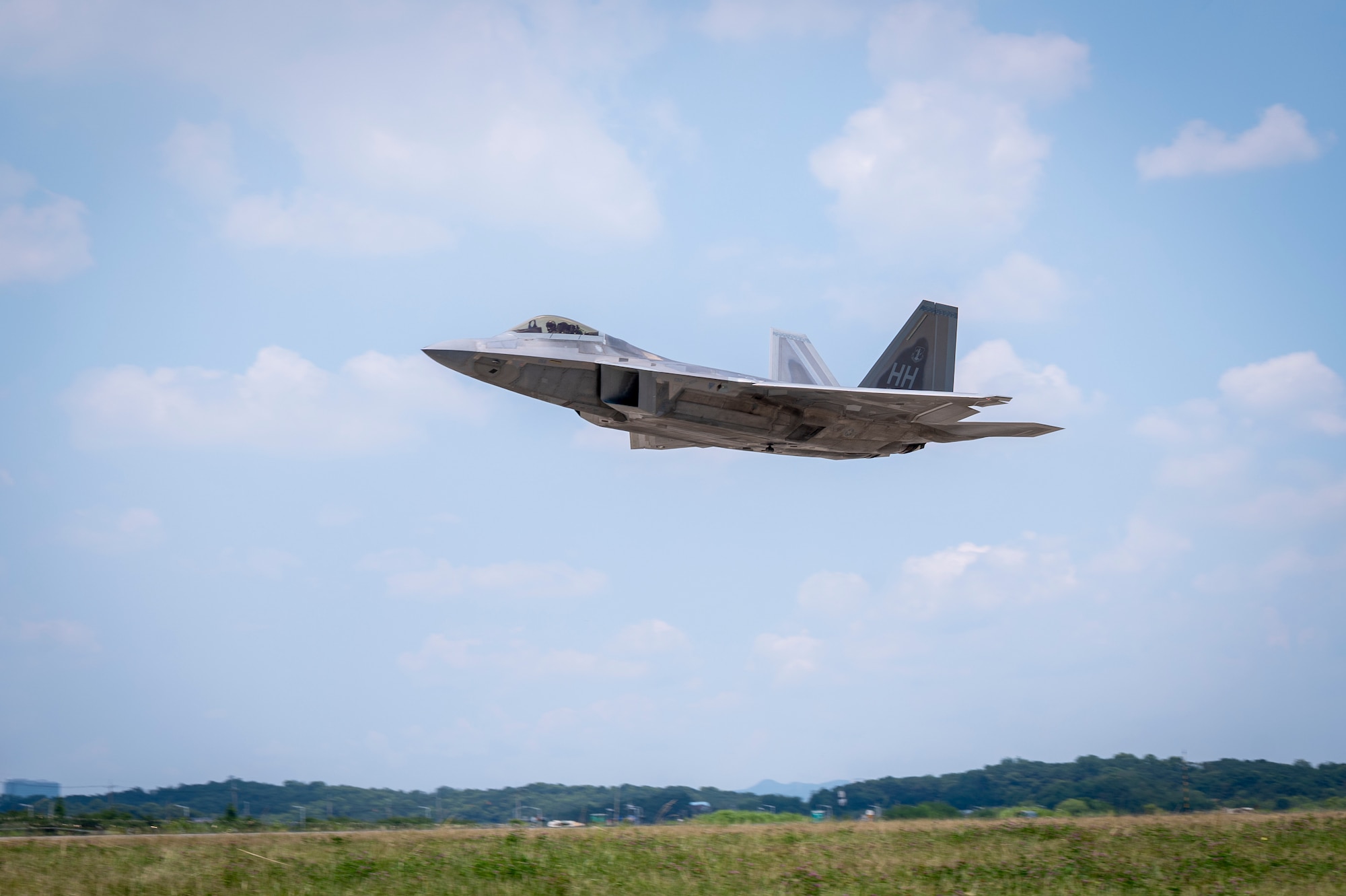 A gray airplane, F-22, appears in a blue sky with small white clouds across the background over a green field with a small dark green line of tree covered hills in the far distance.