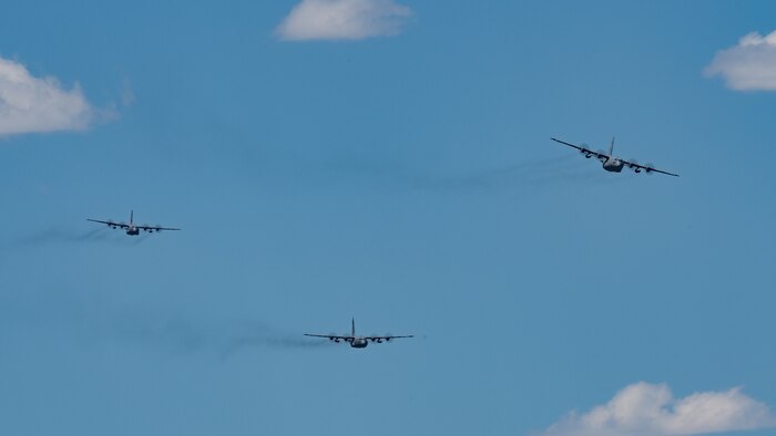 Three C-130s fly in formation against a blue sky in the background.