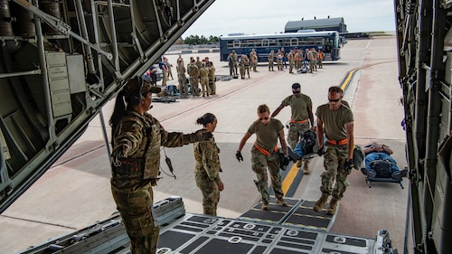 Medics carry a litter into an aircraft with a crowd and blue bus in the background.