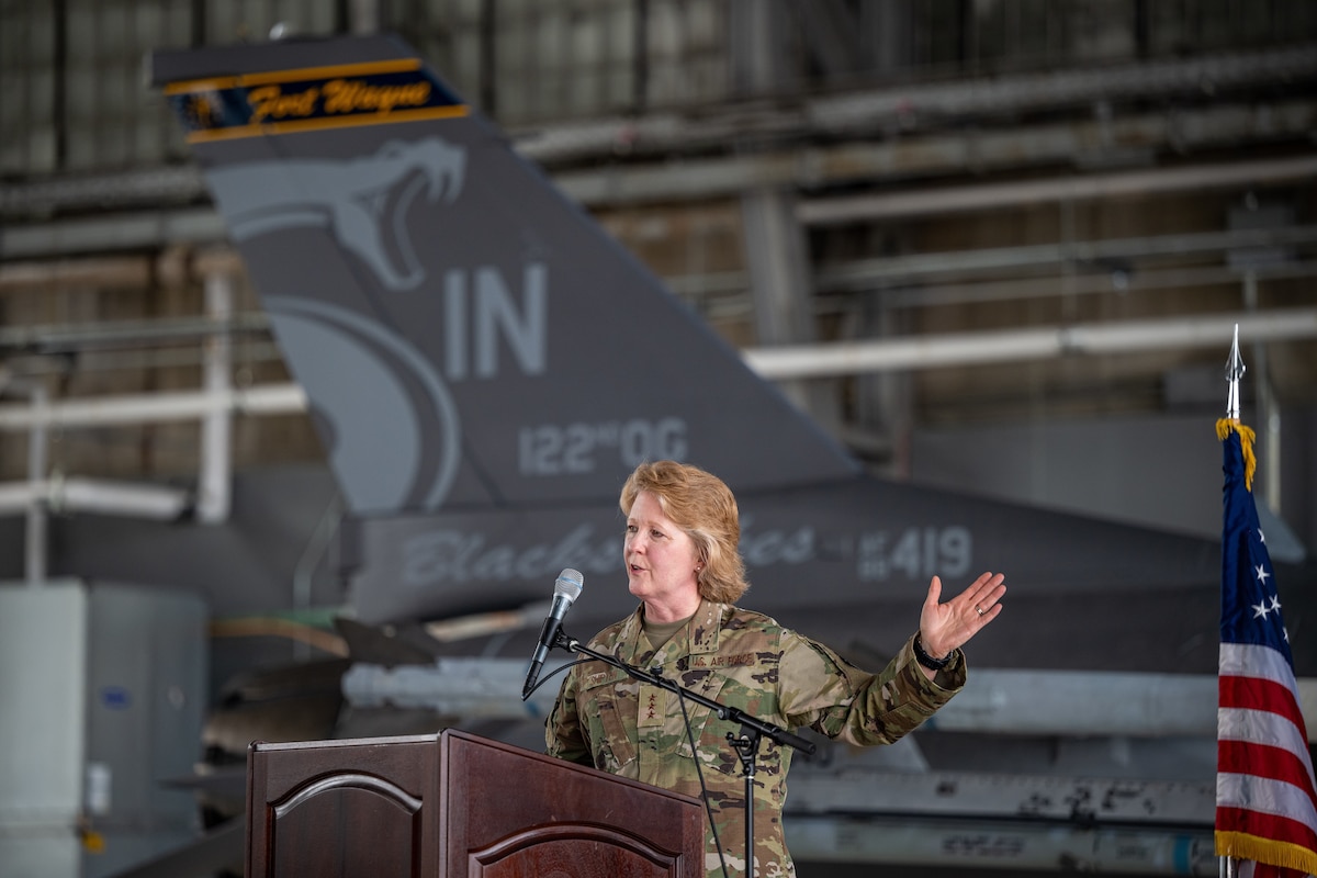 Female general addresses crowd while standing in front of F-16 airframe.