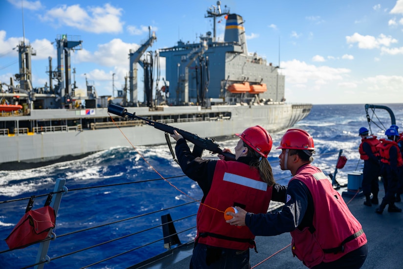 A sailor holding a signal line gun takes aim on a ship takes aim at another ship in the ocean while a second sailor assists from behind.