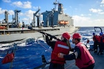 A sailor holding a signal line gun takes aim on a ship takes aim at another ship in the ocean while a second sailor assists from behind.