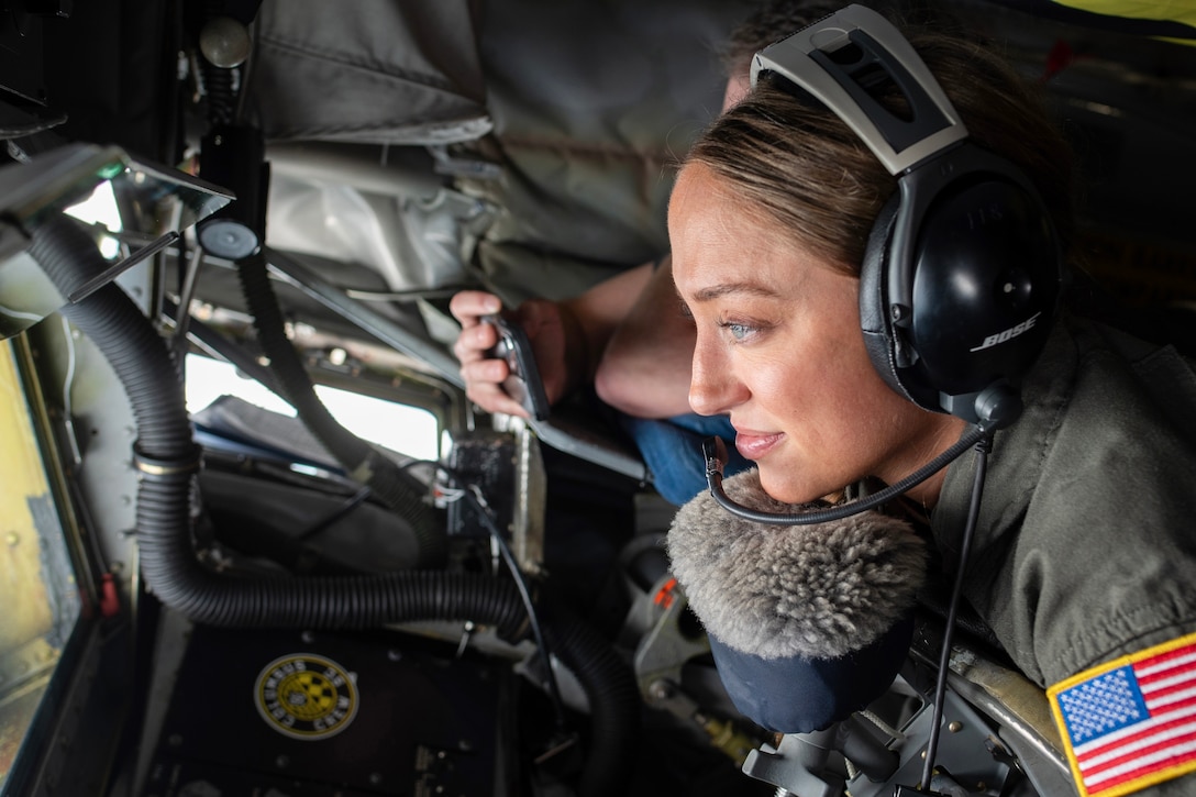 An airman wearing a headset looks out the window of a cockpit; the hand of another person is shown holding a phone.