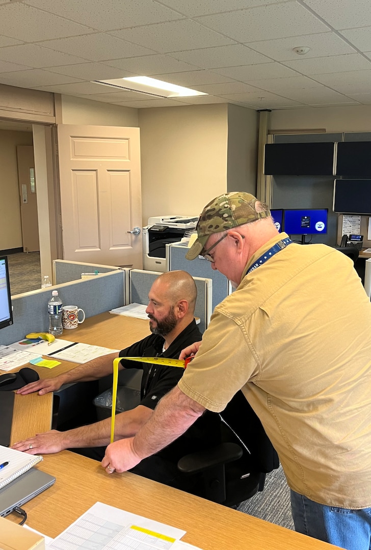 Man reading measuring tap while another man sits in chair at desk.