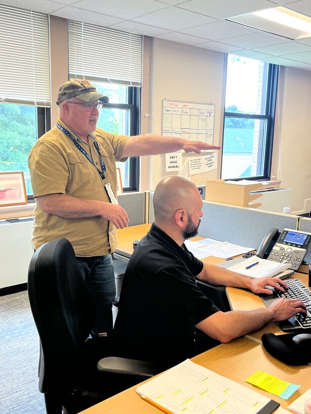 Man standing behind another man who is sitting at desk in  office chair