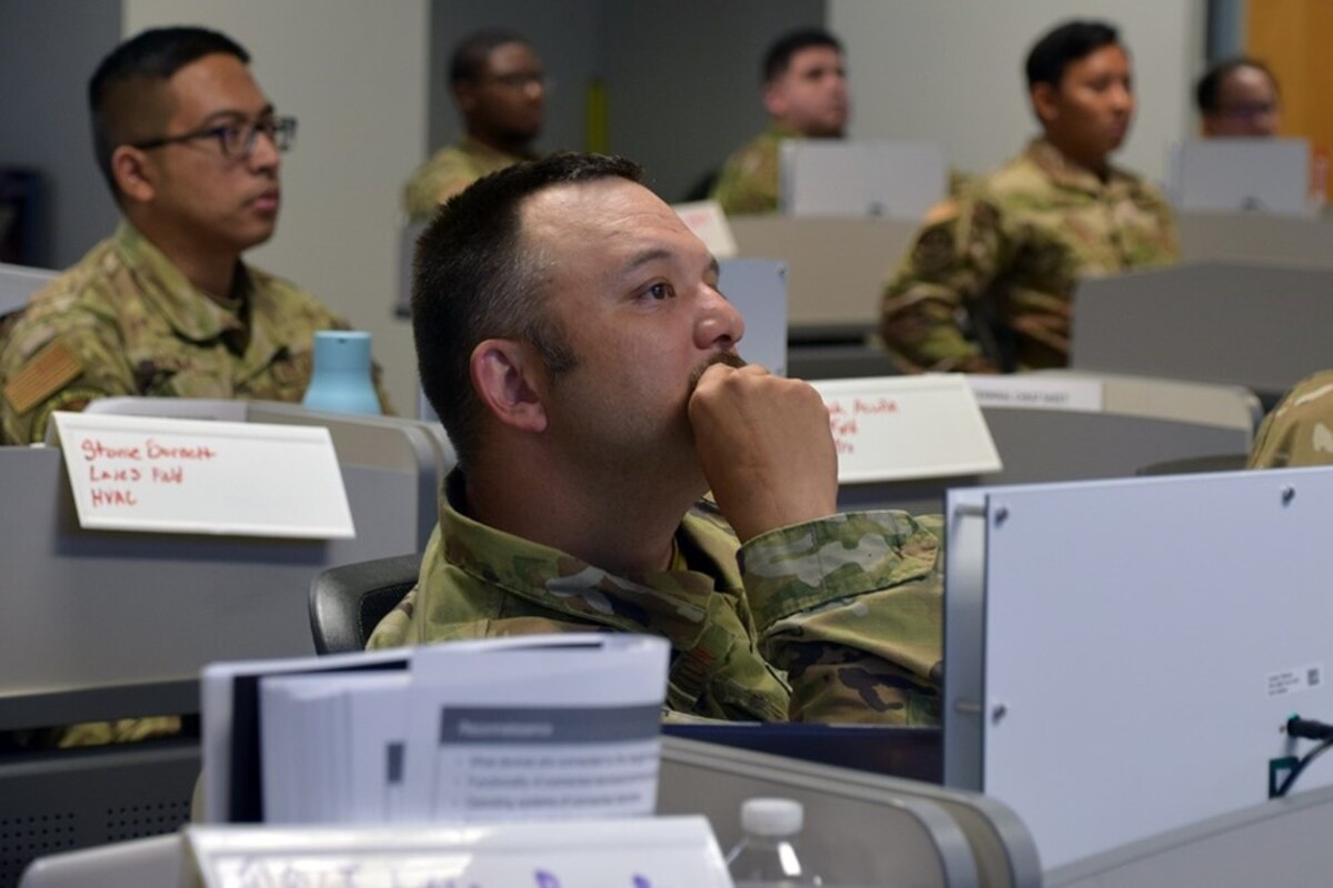 A man in a green uniform sits at a desk during a briefing.