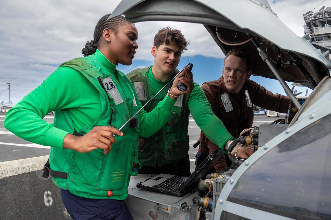 Two sailors standing behind an open section of a helicopter observe a third holding up a small rod with a laptop between them.