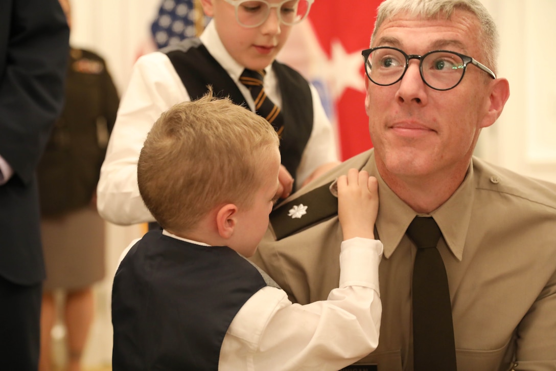 A soldier kneels while two children put a pin on the soldier's shoulder with a flag in the background.