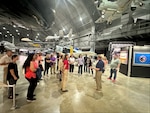 Side view of a man in blue shirt and tan pants speaking to a group of people in an aircraft hangar.