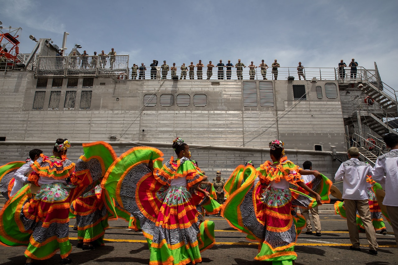 Sailors on top of a Navy ship look at women wearing colorful dresses dance on a pier below.