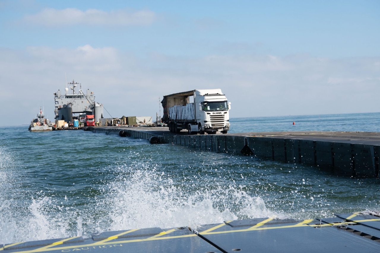 A large truck rolls across a narrow pier as waves lap against a structure in the foreground.