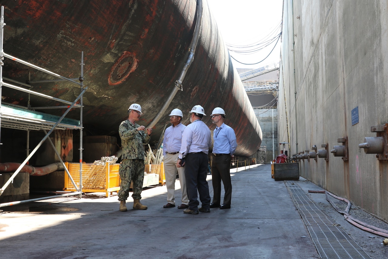 Men wearing hard hats stand near the hull of a submarine in dry dock.
