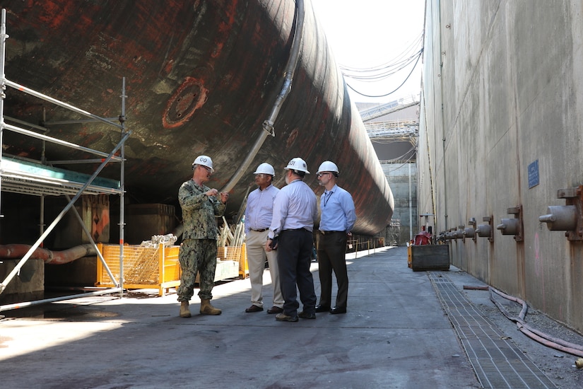 Men wearing hard hats stand near the hull of a submarine in dry dock.