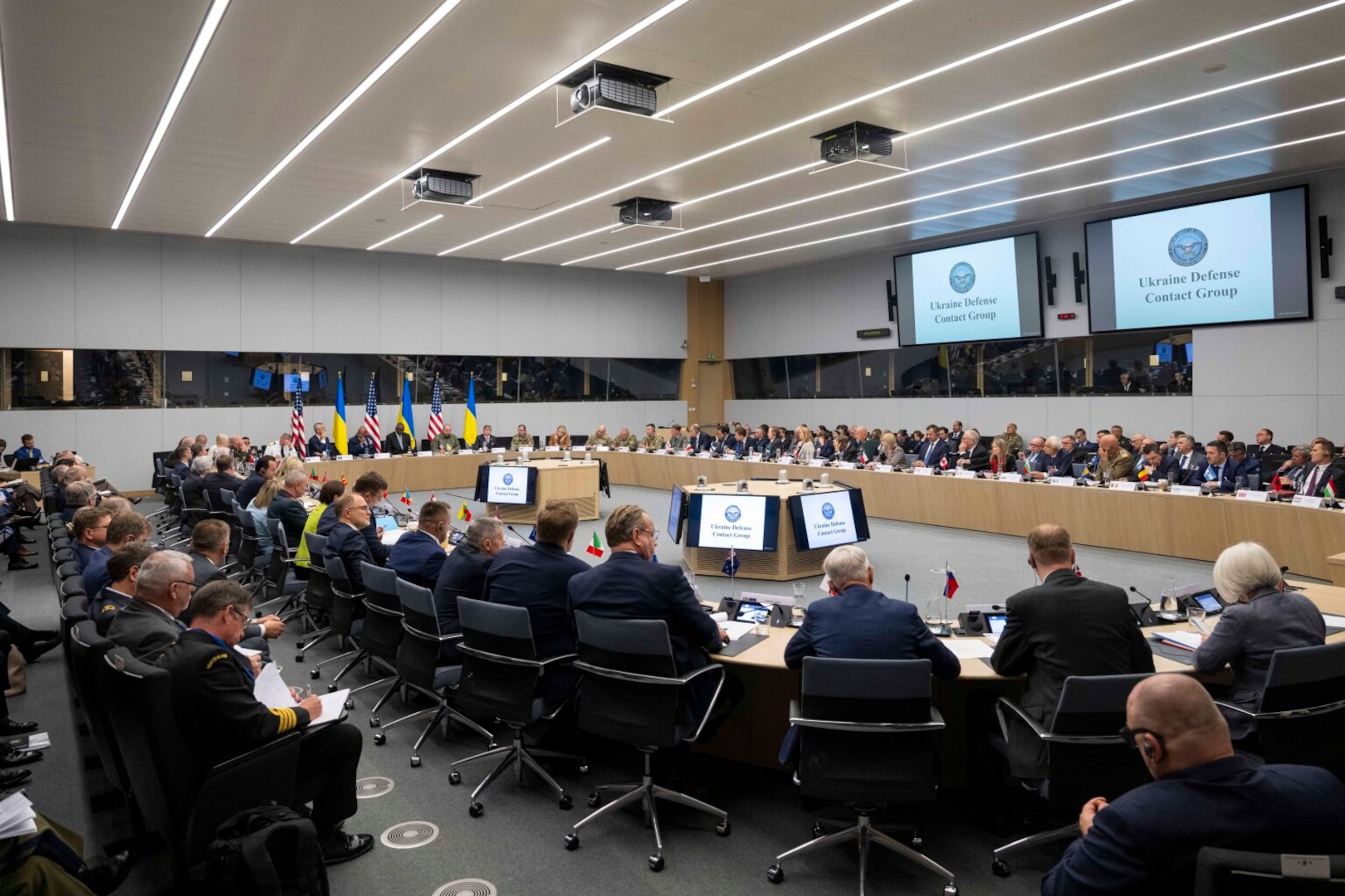 A group sits around a large oval table with screens displaying a slide that reads “Ukraine Defense Contact Group.”