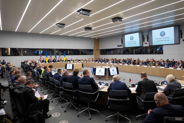 A group sits around a large oval table with screens displaying a slide that reads “Ukraine Defense Contact Group.”