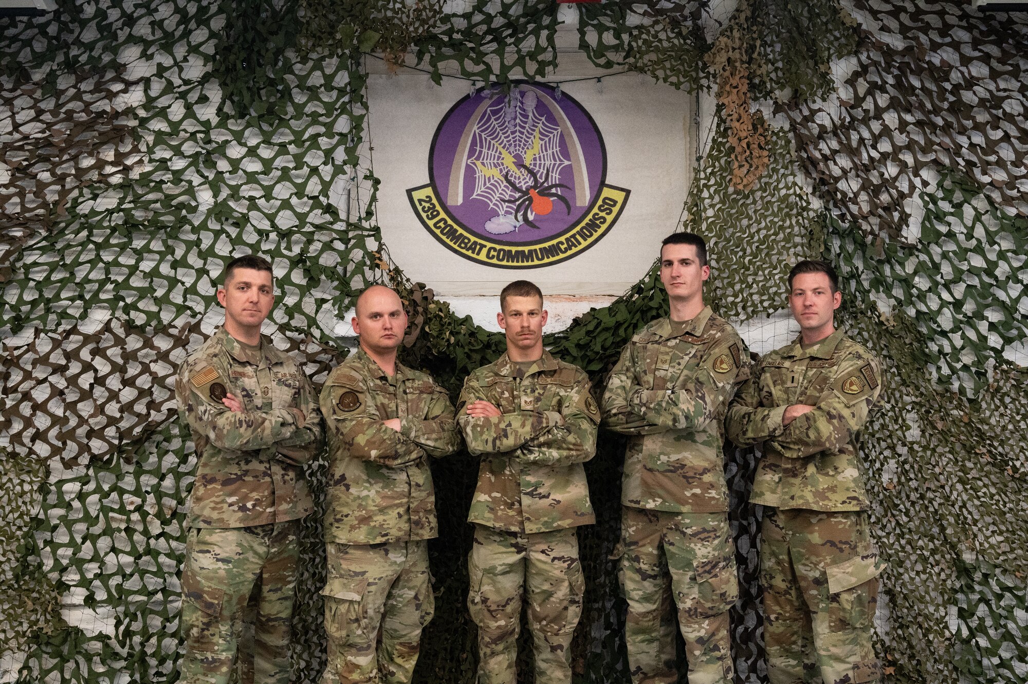U.S. Air National Guard Airmen pose for a photo in front of their unit emblem and mosquito netting. The Airmen are wearing OCP pattern uniforms.