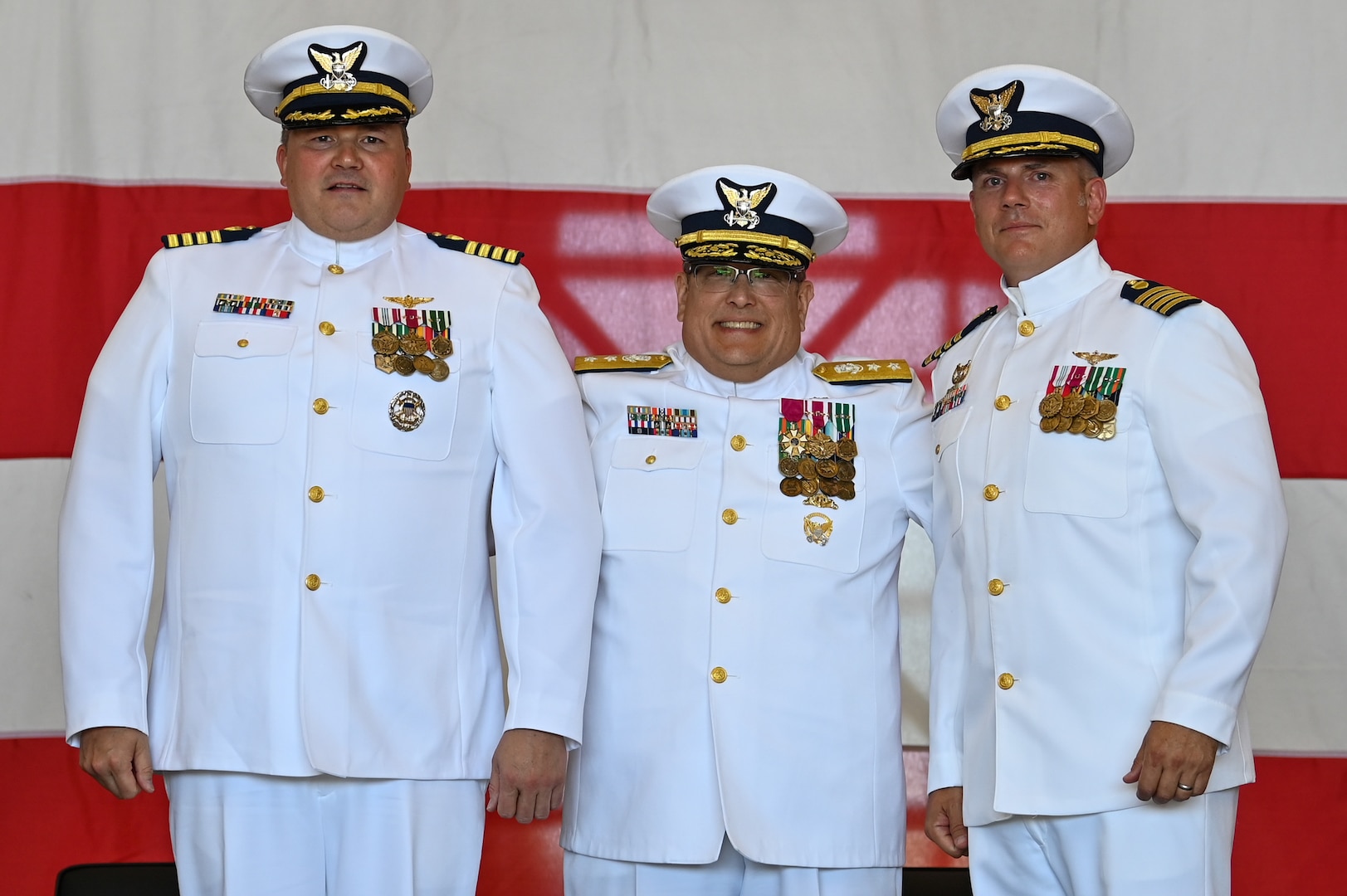 3 U.S. Coast Guard officers are seen standing in front of a large American flag