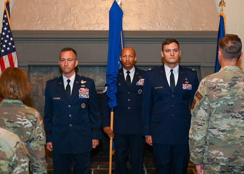 Three airmen in uniform stand facing an audience and holding a flag, with two flags on either side.