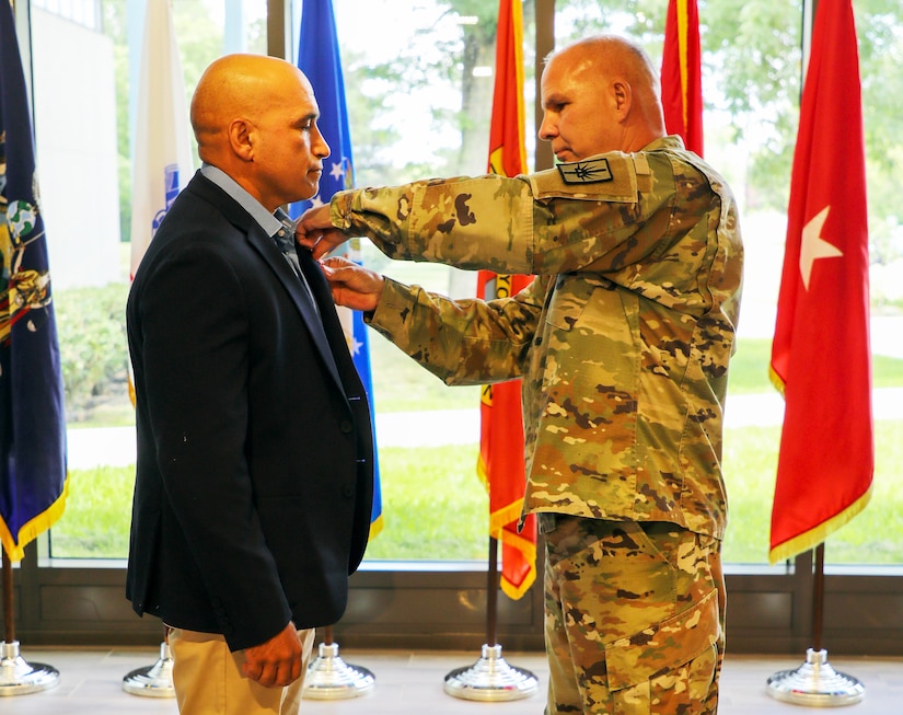 A service member pins a medal on a veteran in front of five flags and a window in the background.