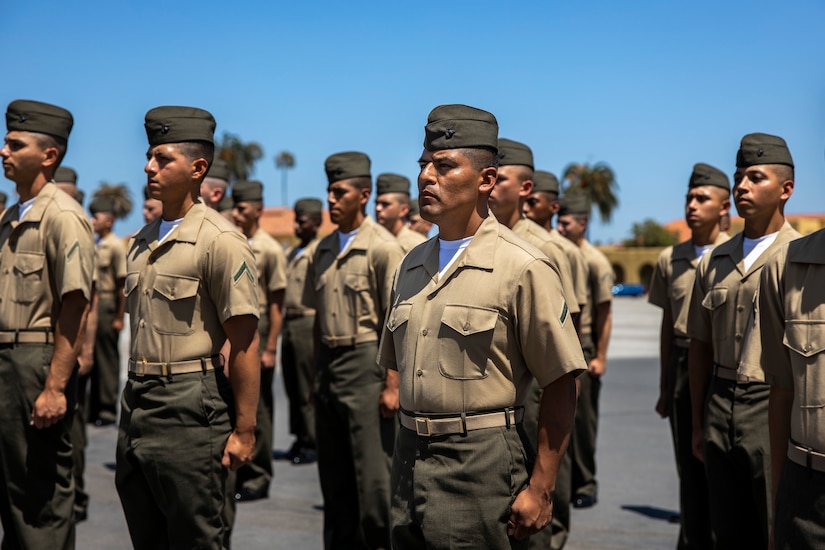 A group of soldiers in uniform stand in formation facing left on a paved surface on a clear day.