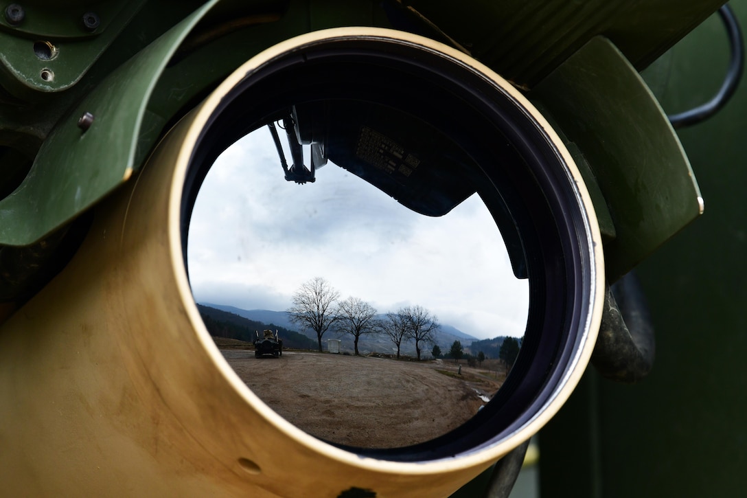 A close-up of the large circular lens and protective shield on a mounted weapon displaying a wide field with trees on a cloudy day.