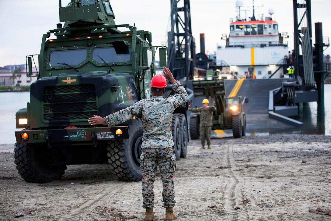 A Marine wearing a red helmet signals with both hands to large vehicles driving off a water vessel at a dock.