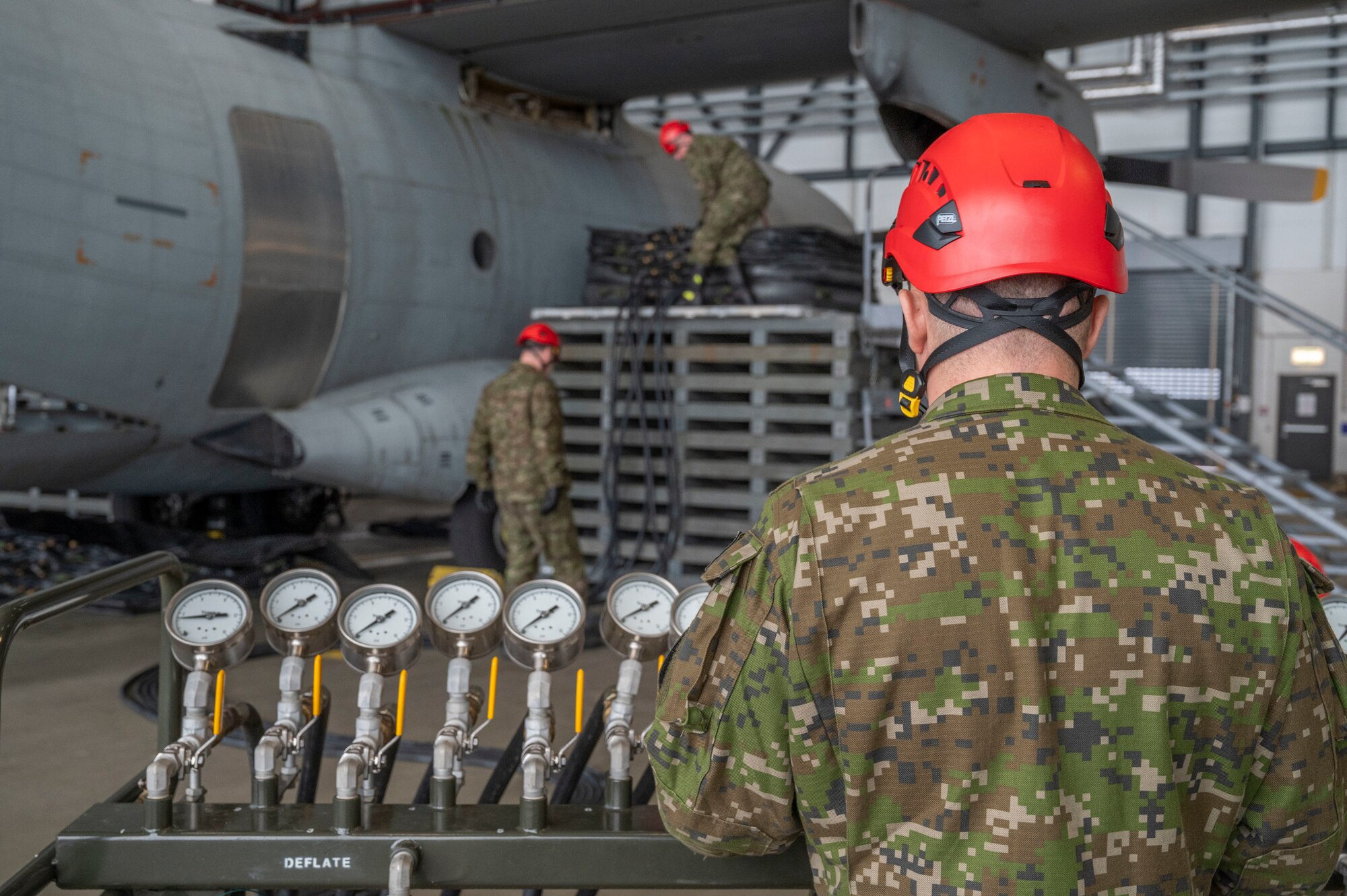 A man stands in front of an air manifold console, two men are behind him adjusting hoses for an airbag that is about to be used to lift a plane by its wings.
