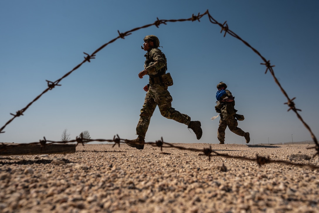 Two airmen run across gravel and sand with barbed wire in the foreground.