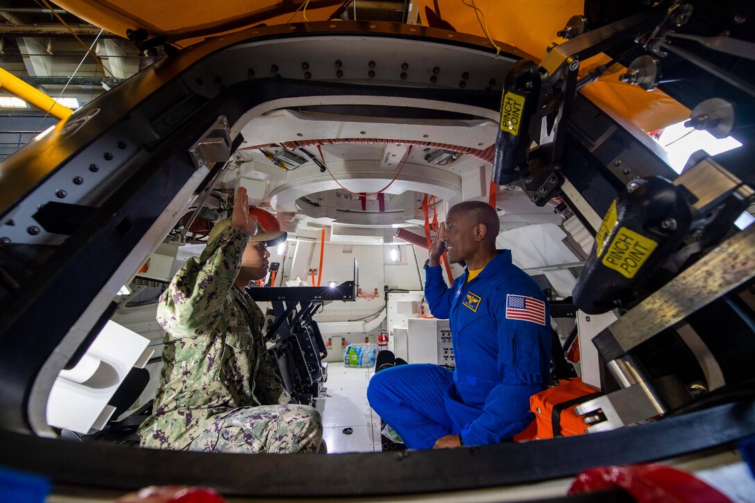 Two sailors raise their right hands as they look at each other in a space capsule.