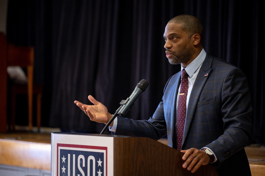 A man in a coat and tie gestures as he speaks at a podium with a USO sign on it.