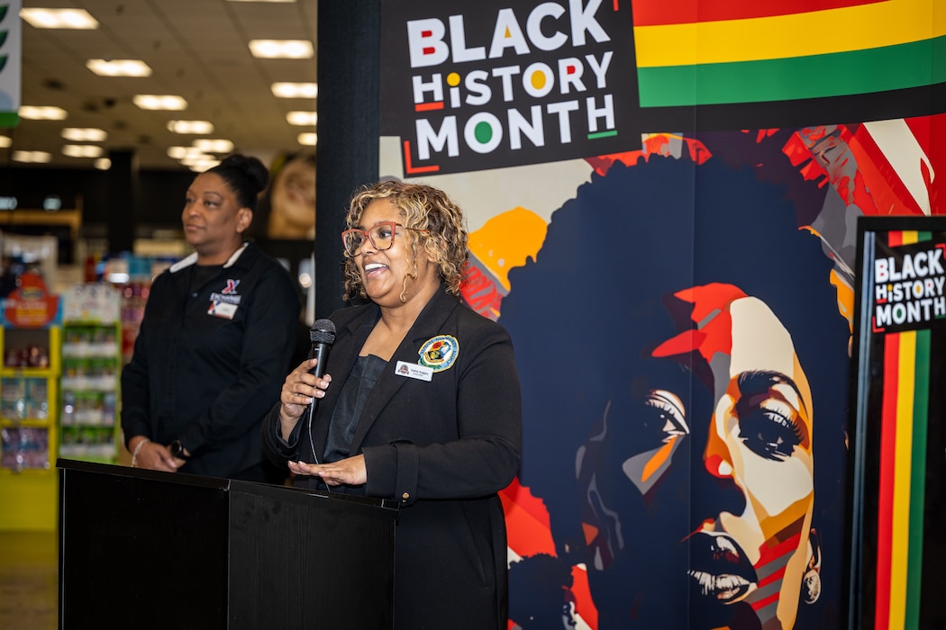 A woman speaks at a podium. Behind her is a graphic sign that shows a stylized African-American face and the words "Black History Month". To the left is the entrance to the base exchange store.