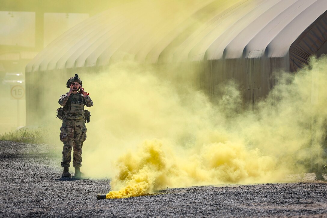 A solider standing on gravel next to a tent shouts as clouds of yellow smoke fill the air.
