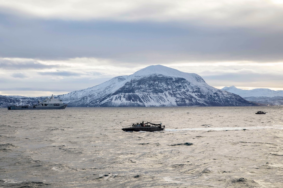 Three boats transit a body of water under a cloudy sky with snowy mountains in the background.