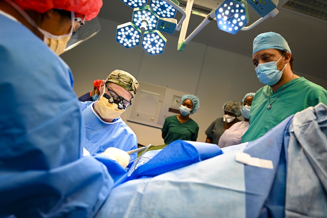 Spotlights shine as doctors perform a surgery while medical team members watch in the background.