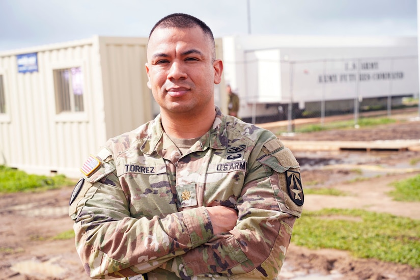 A service member looks into the camera with his arms crossed; behind him is a grassy area with portable buildings.