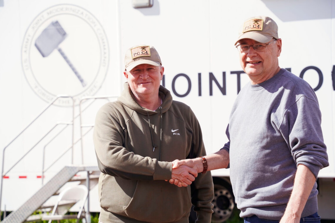 Two men dressed in civilian clothing pose for a photo while shaking hands. Behind them is a portable building.