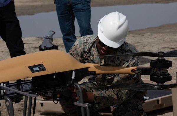 A Marine in uniform wearing a hard hat examines a small, unmanned aircraft.