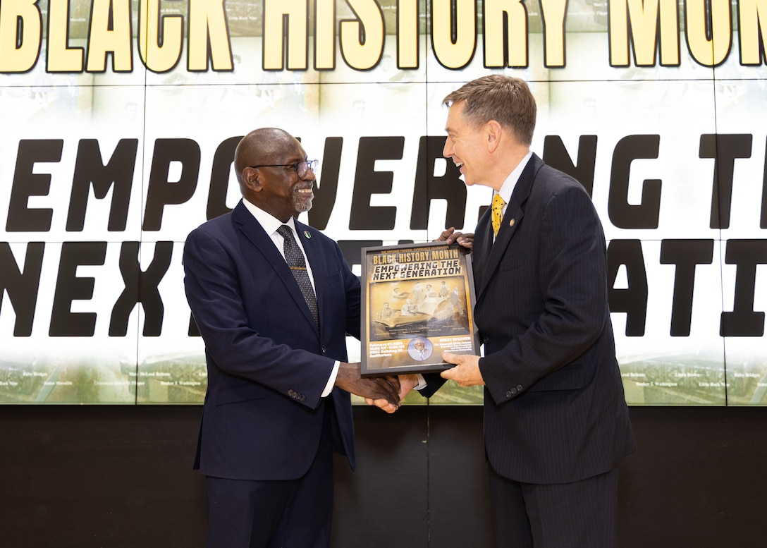 Two men in dark suits hold a memento and shake hands. One is dark skinned and bald and the other light skinned with graying hair.