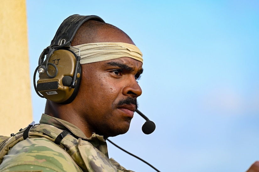 A soldier wearing a headband and headset looks into the distance with a blue sky in the backdrop.